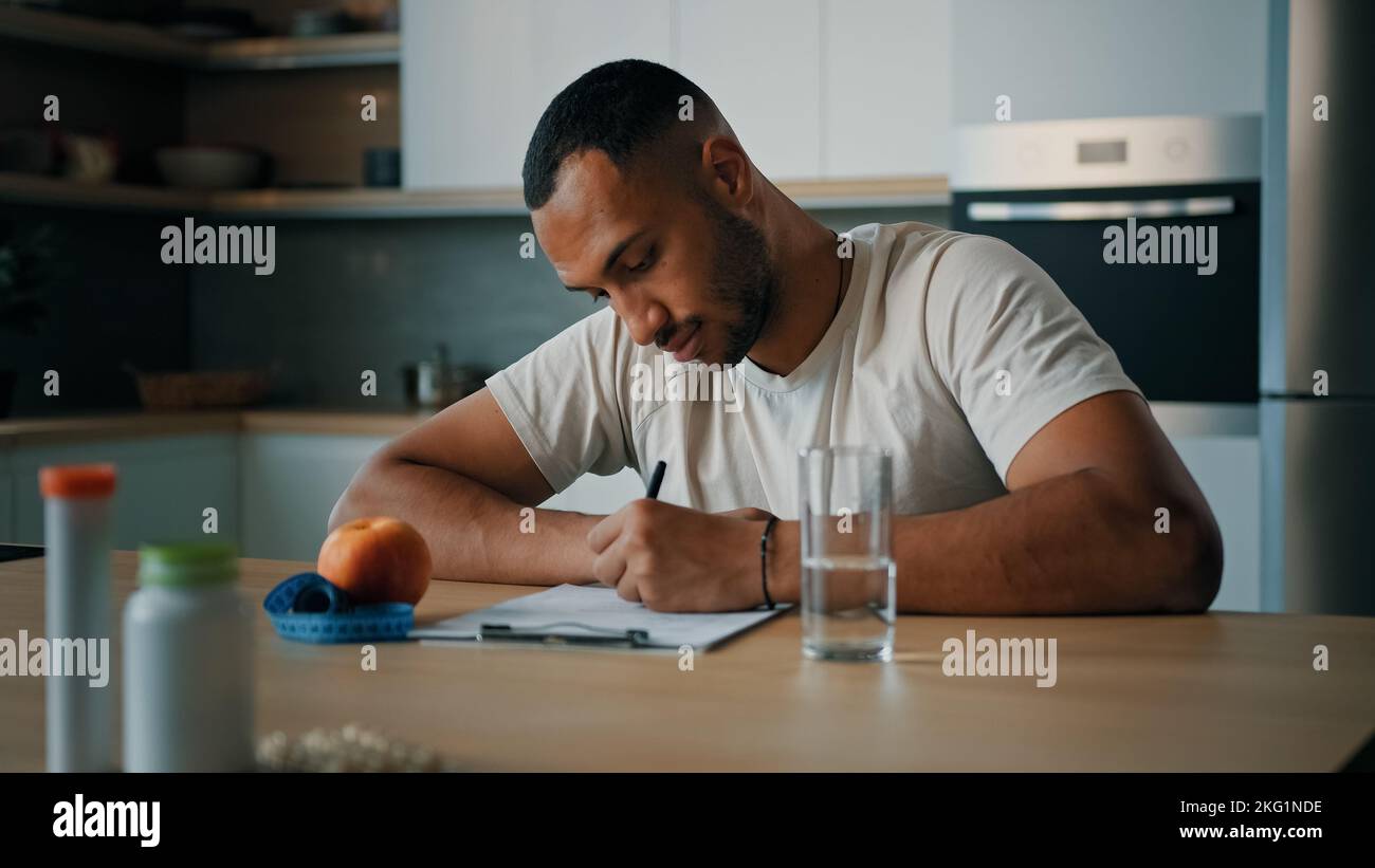 African American man guy male sports athlete nutritionist specialist sitting at table in kitchen writing prescription sport fitness diet on paper Stock Photo