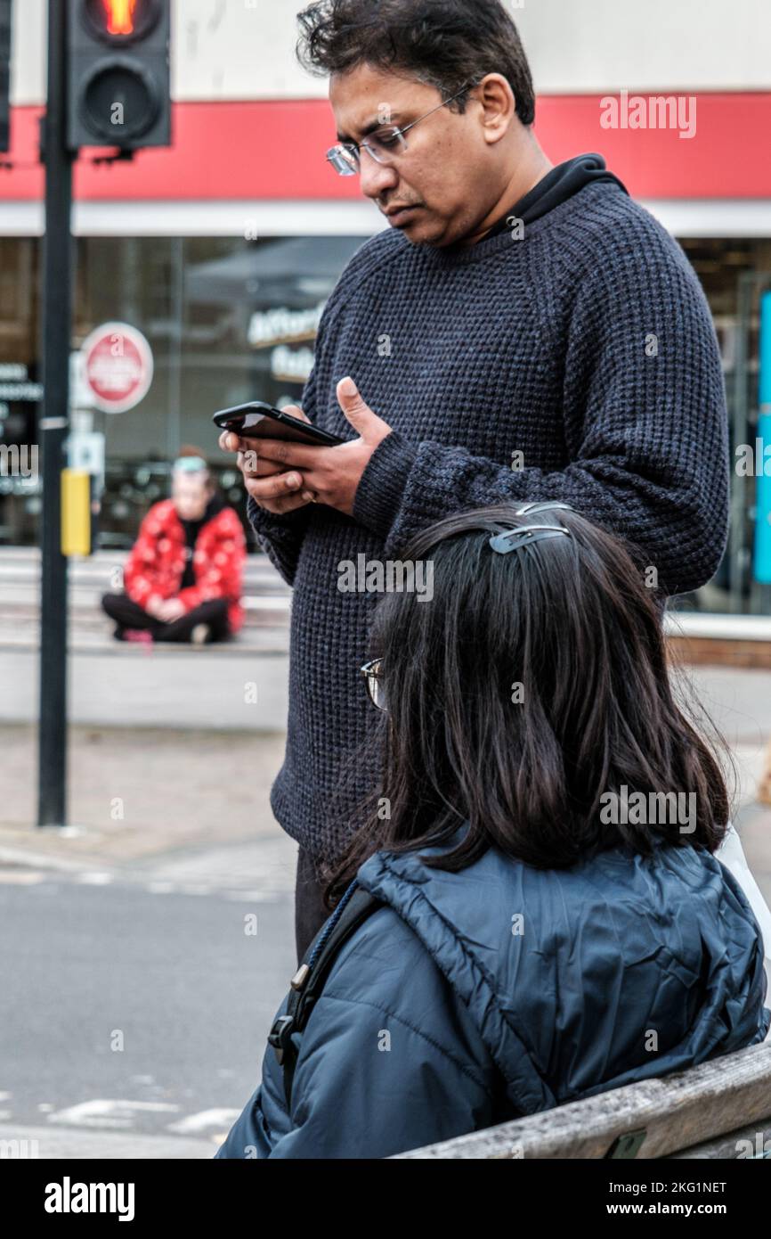 Epsom, Surrey, London UK, November 19 2022, Asian Ethnic Couple Man Standing Using A Mobile Phone Or Smartphone Woman Stting On A High Street Bench Stock Photo