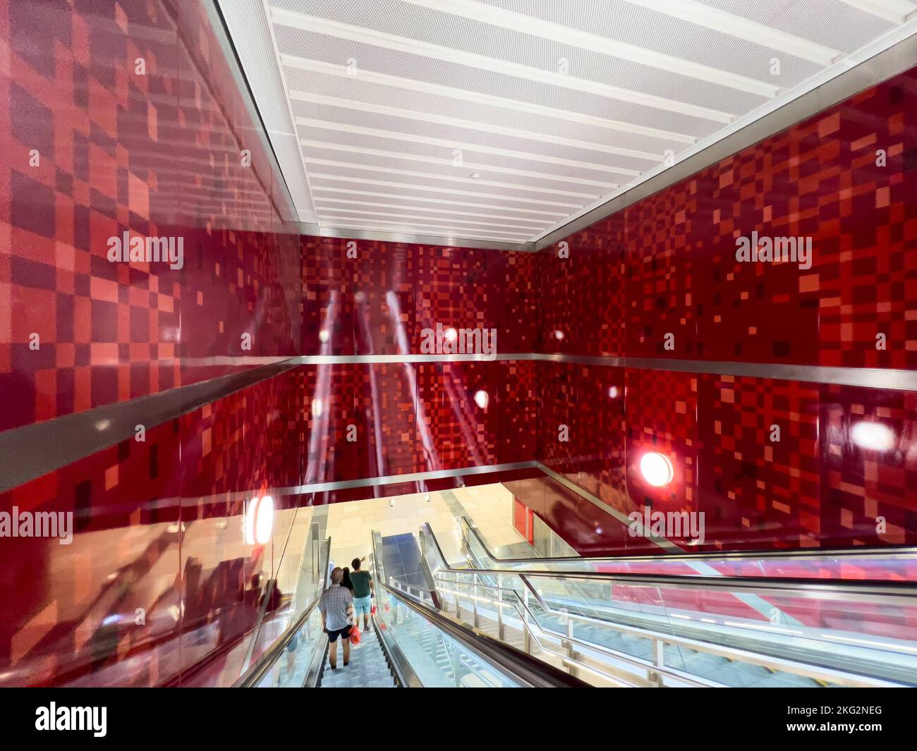 Commuters travel deep down a long escalator within a red colour wall interior design. Stock Photo