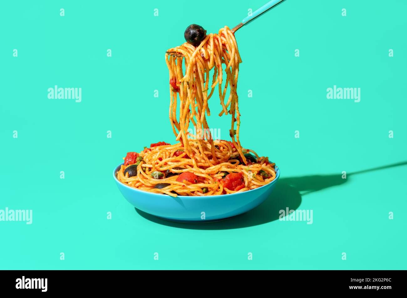 Taking pasta with a fork from a bowl, minimalist on a green table. Vegan Italian dish, spaghetti puttanesca, with tomato sauce, black olives, and cape Stock Photo