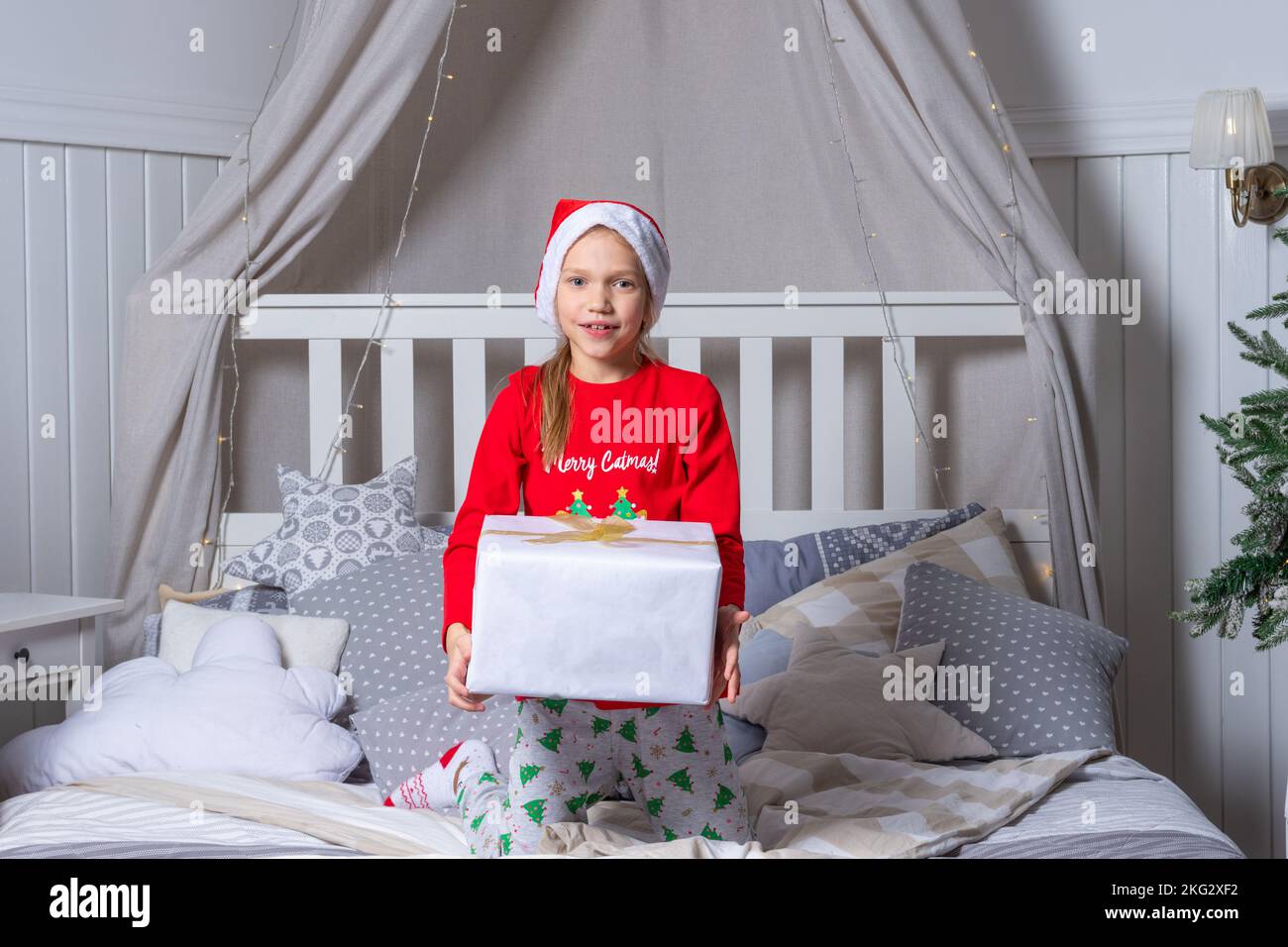 Happy funny baby girl in pajamas and Santa hat holding a gift box in bed on Christmas morning. Children are enjoying the Christmas holiday. The child Stock Photo
