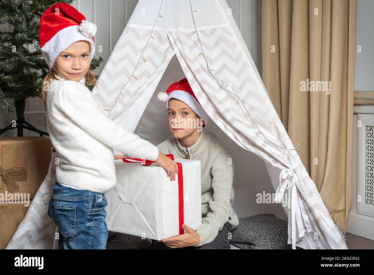 A girl in a Santa hat gives a gift to a boy for Christmas and New Year in the nursery. Brother and Sister exchanging gifts. Children give gifts. Merry Stock Photo