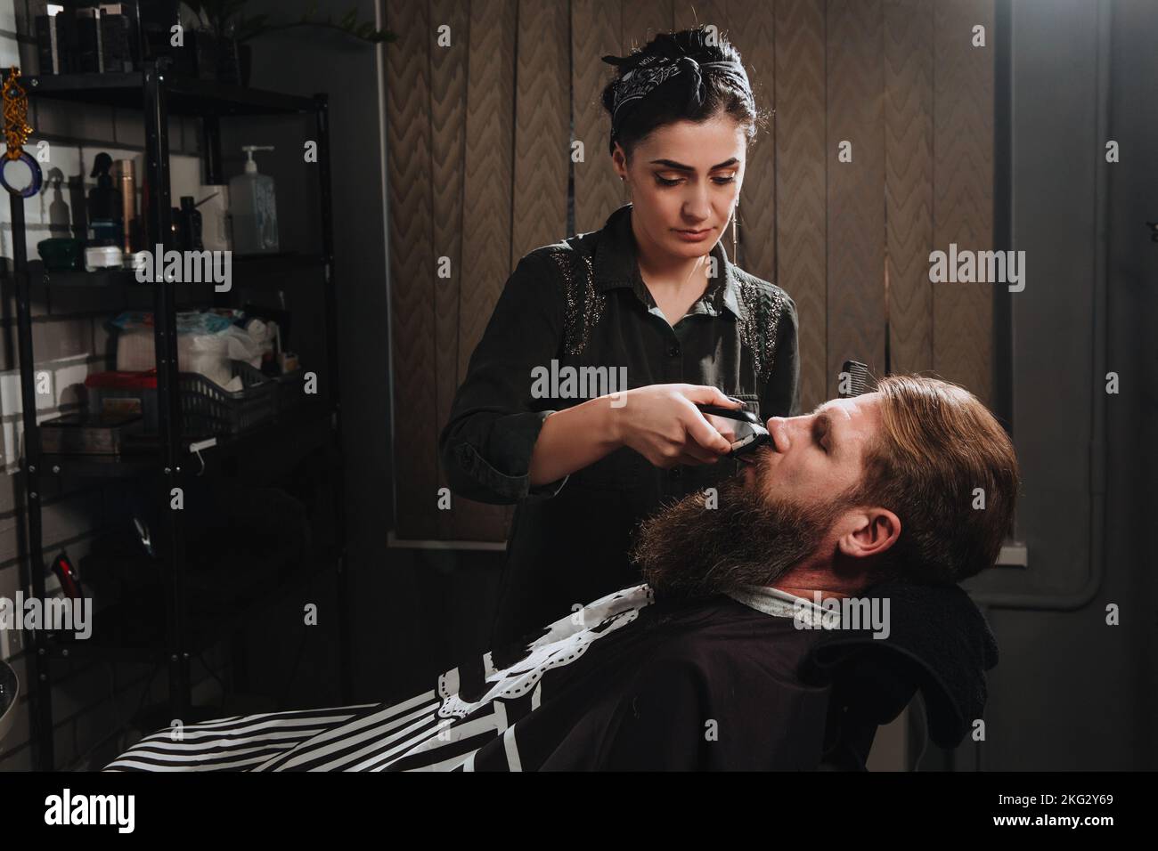 Young woman barber making haircut of bearded man in barbershop. Self-care, masculine beauty. Hair care service concept Stock Photo