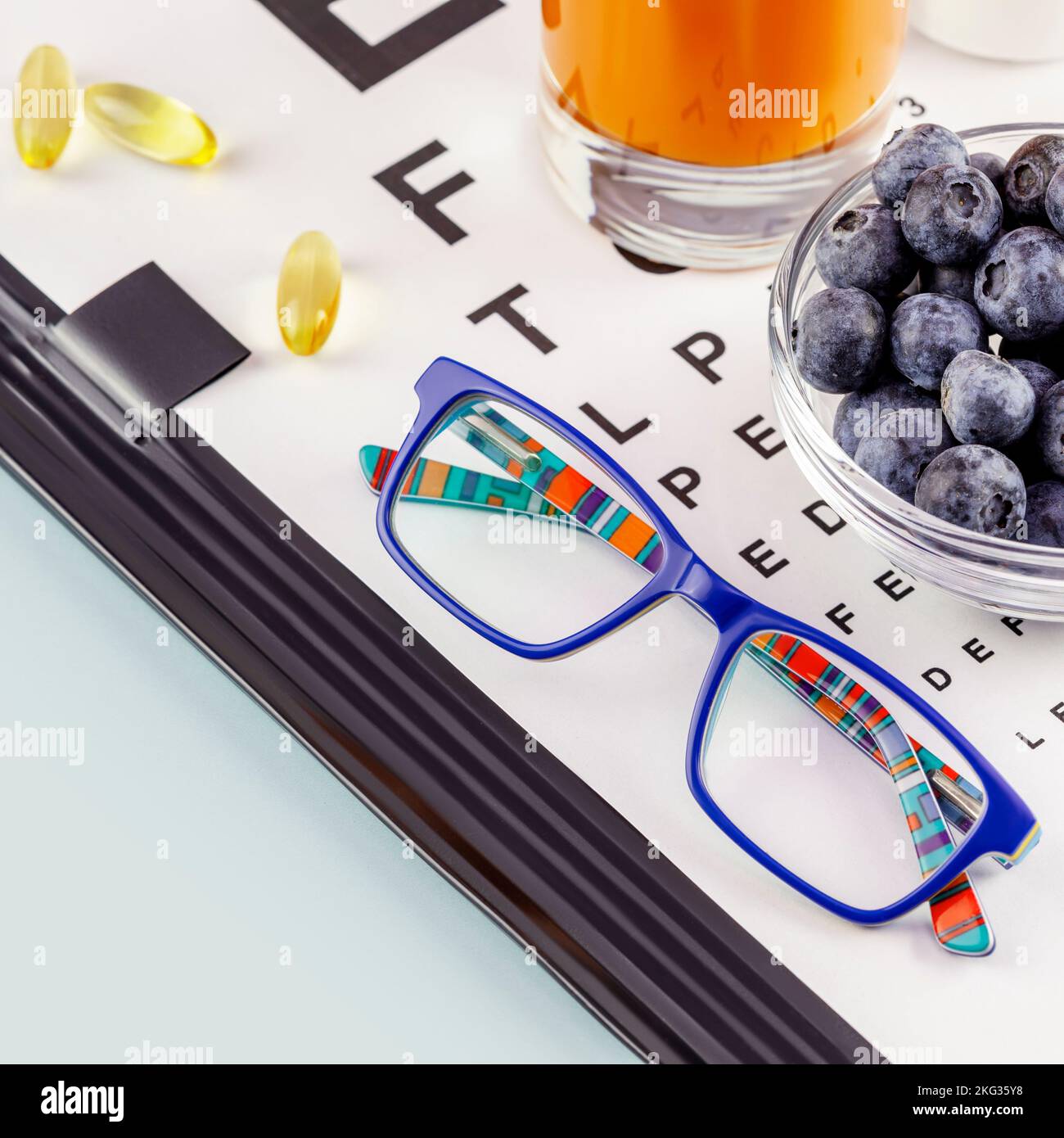 Kid eyeglasses, glass of carrot juice, bowl with blueberries and Snellen table for vision testing on blue background. Nutritions for good vision. Vita Stock Photo