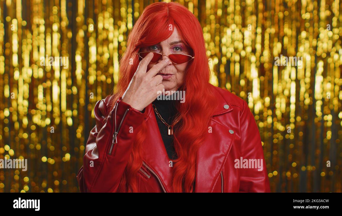 I am watching you. Woman with red hairstyle wig pointing at his eyes and camera, show I am watching you gesture, spying on someone. Senior elderly grandmother isolated alone on shiny studio background Stock Photo