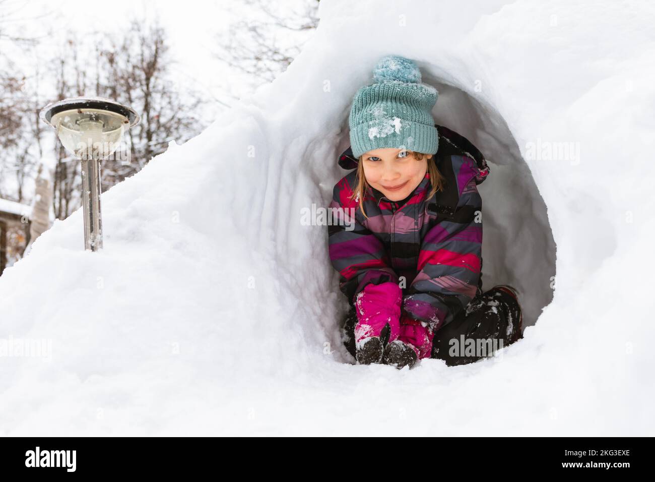 Happy little girl sitting in a snow cave made in a snowdrift looking at camera smiling Stock Photo
