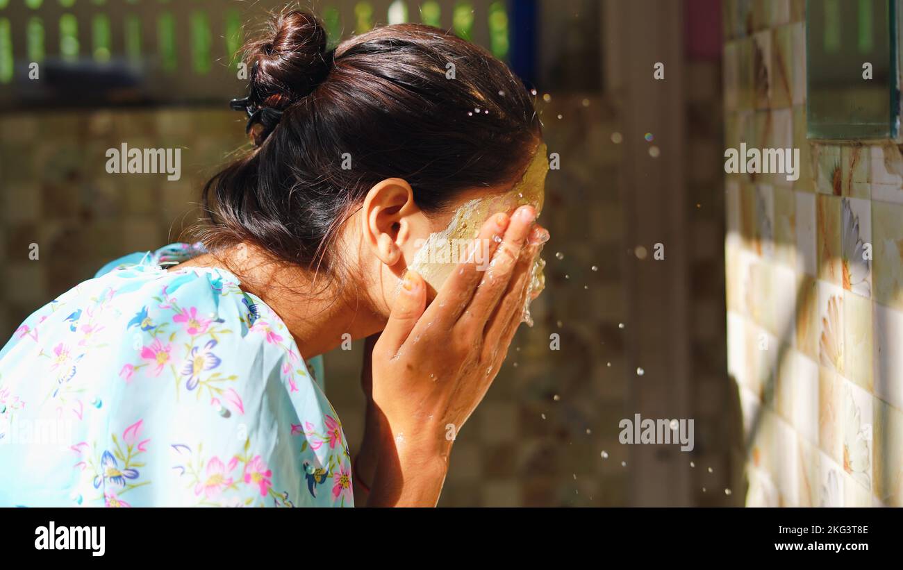 Beautiful woman is washing facial mask in bathroom after applying face mask. Girl spraying water on her face standing in front of mirror at home. mud Stock Photo