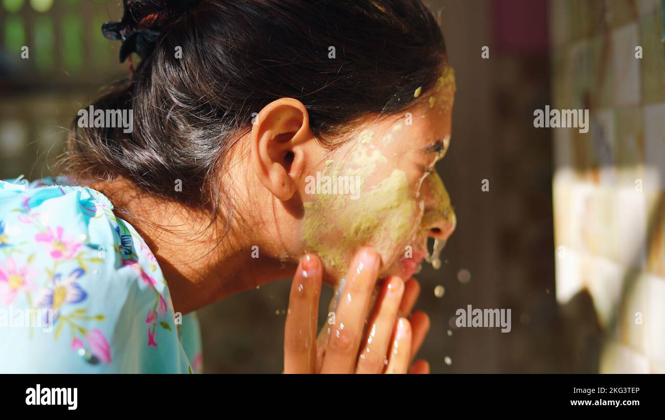 Beautiful woman is washing facial mask in bathroom after applying face mask. Girl spraying water on her face standing in front of mirror at home. mud Stock Photo