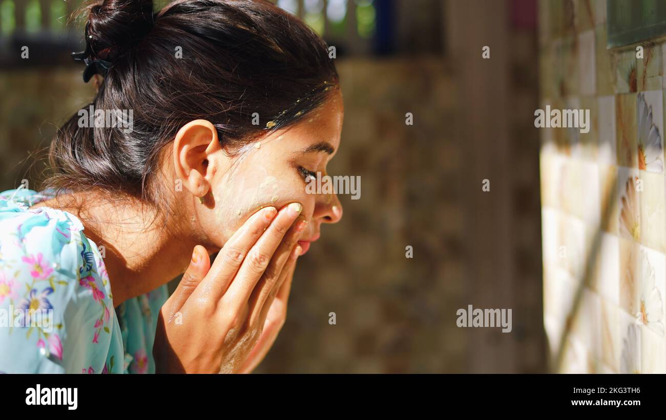 A beautiful woman is washing facial mask in bathroom after applying face mask. Girl spraying water on her face standing in front of mirror at home. mu Stock Photo