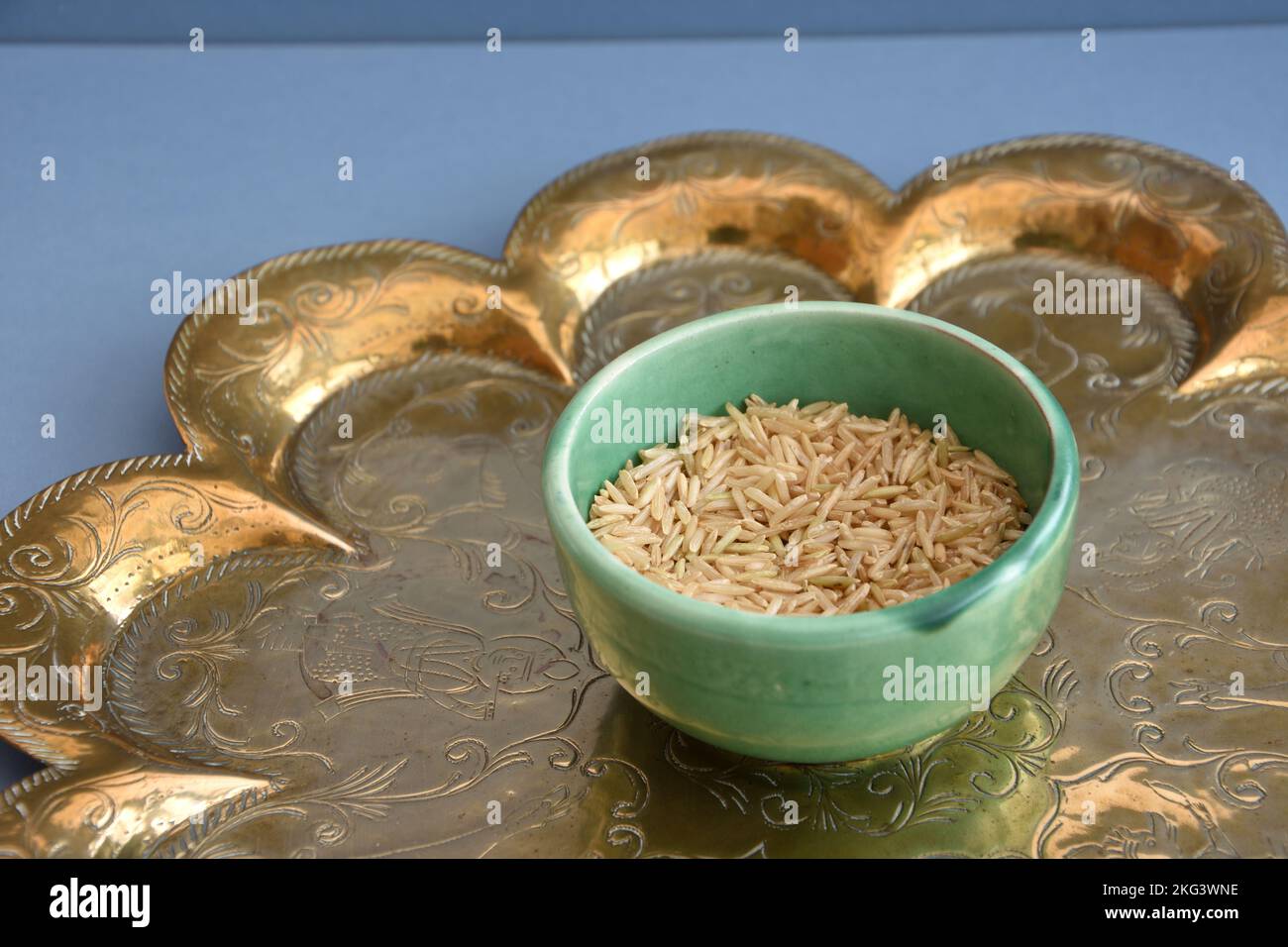 Basmati brown uncooked rice in green ceramic Brannam bowl on an antique Indian brass tray on a blue background. Weighed in preparation for cooking a c Stock Photo