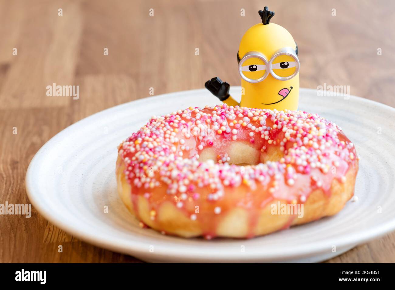 A toy model of The Minion character Kevin looks down on a home made Air Fryer baked glazed doughnut. The doughnut has a pink glaze and sprinkles Stock Photo
