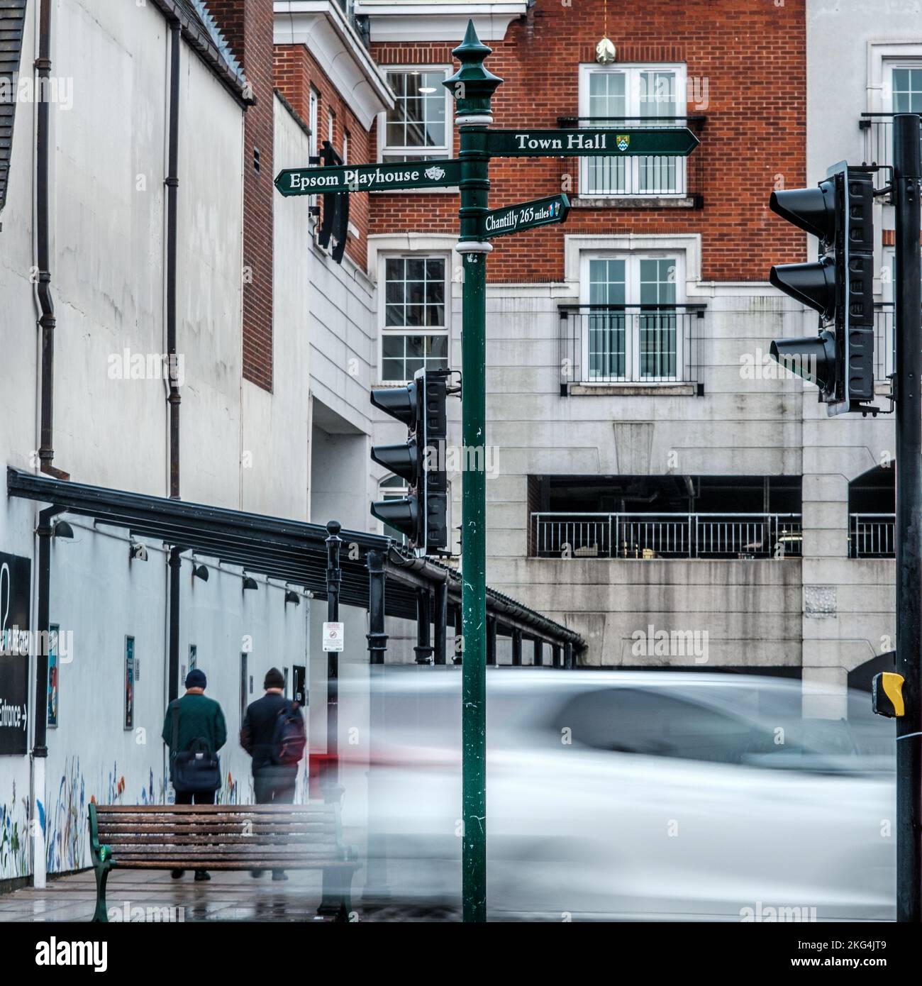 Epsom, Surrey, London UK, November 20 2022, Speeding Blurred Car With Two Men Walking Backs To The Camera Towards A Modern Town Centre Building Stock Photo