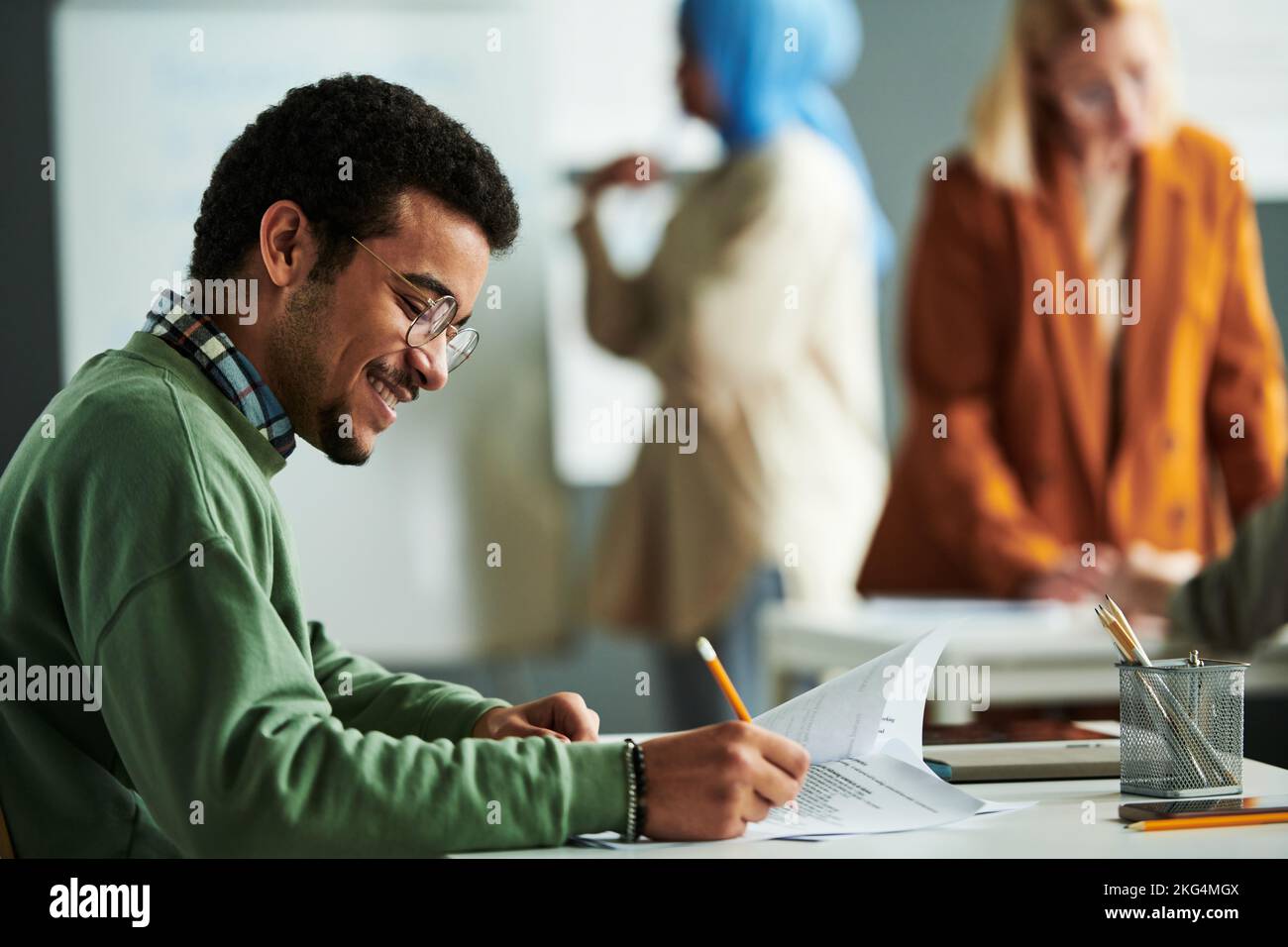 Side view of happy Middle Eastern student putting ticks in front of right answers while carrying out grammar test by desk in classroom Stock Photo