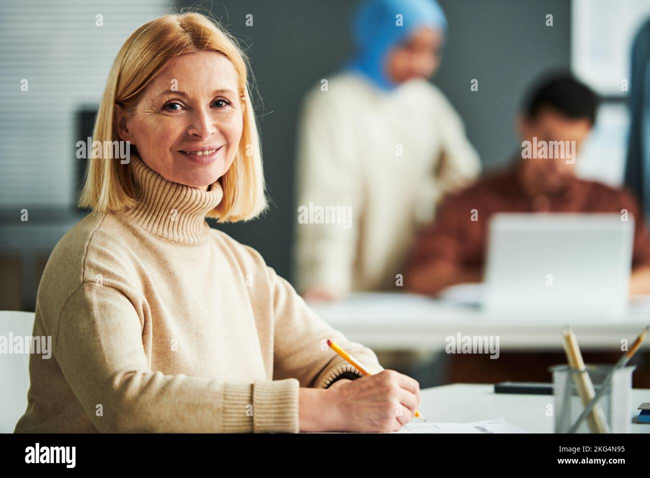 Successful experienced teacher with blond hair looking at camera while sitting by desk in classroom and checking exam tests of students Stock Photo