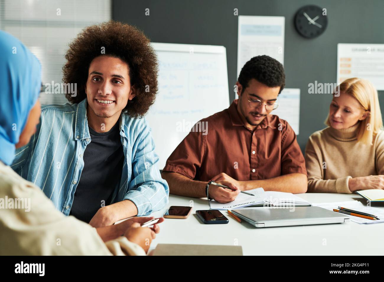Happy young man with thick dark hair looking at Muslim female colleague in blue hijab during conversation by workplace Stock Photo