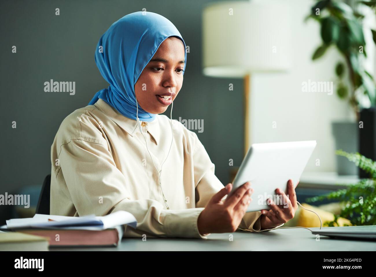 Confident Muslim female teacher or student in blue hijab looking at online audience on tablet screen while communicating to them Stock Photo