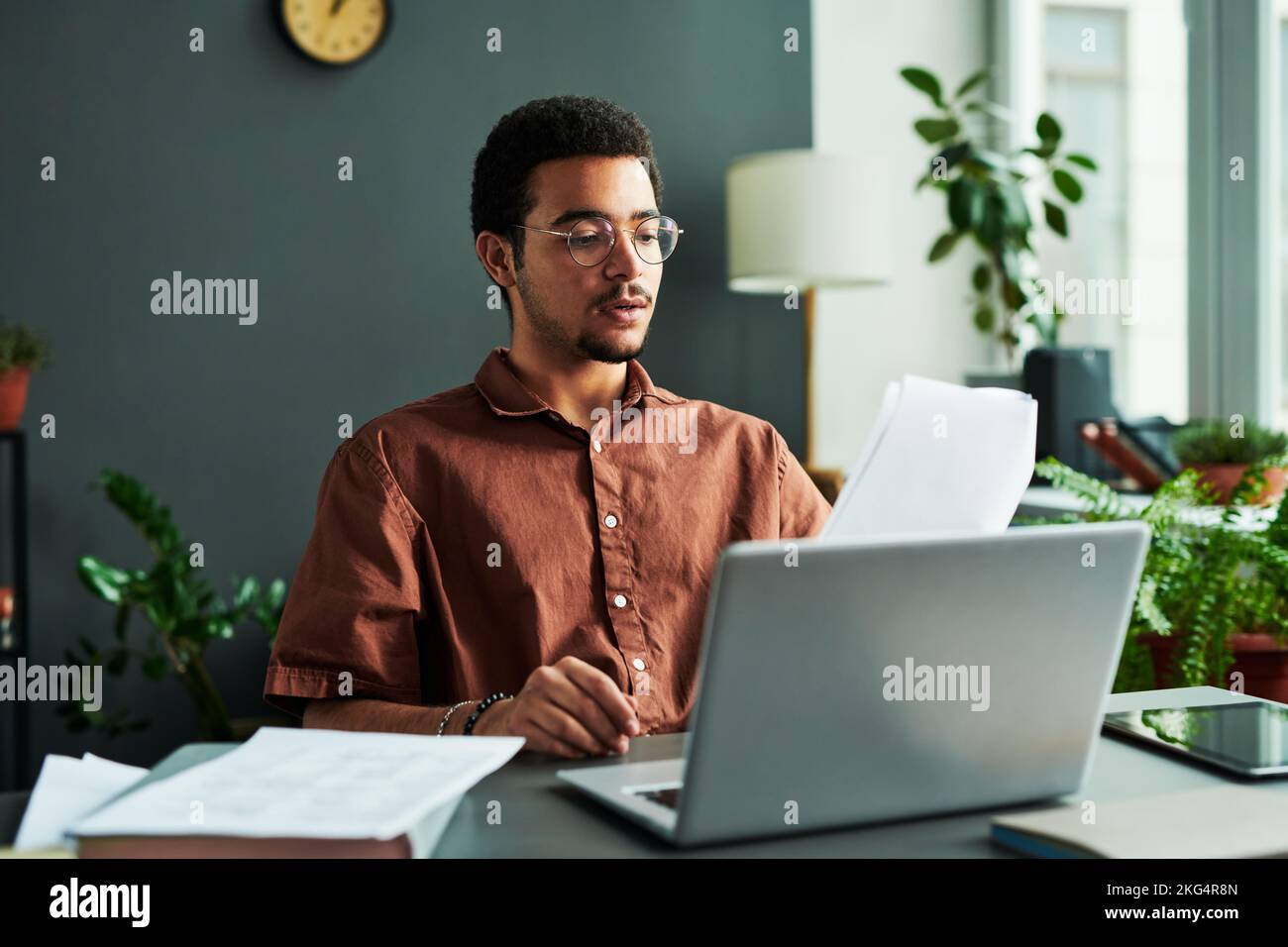 Young confident man with papers sitting by workplace in front of laptop and making report or explaining new information to online audience Stock Photo