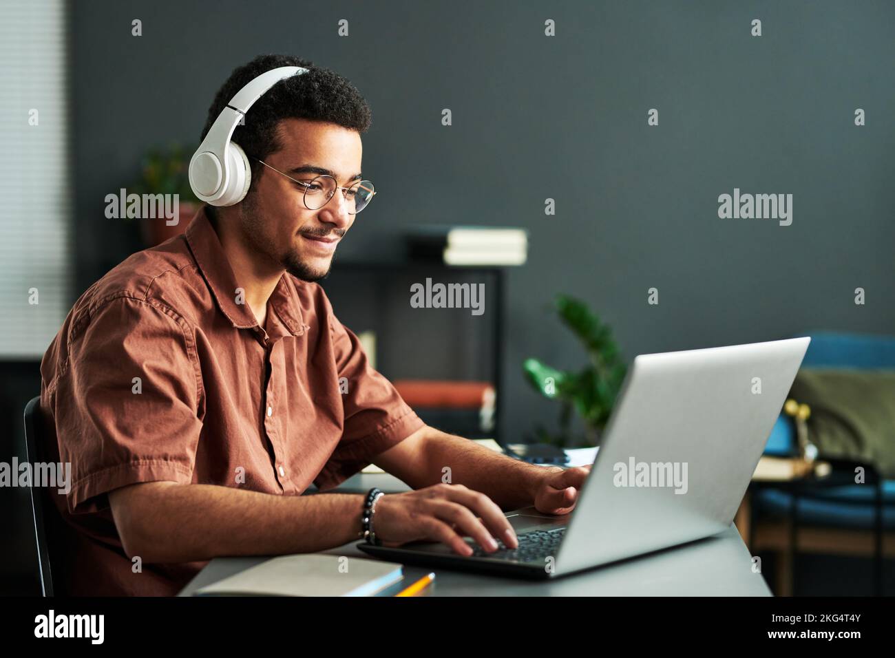 Young smiling man in headphones typing on laptop keyboard while sitting by workplace and taking part in online webinar or lesson Stock Photo