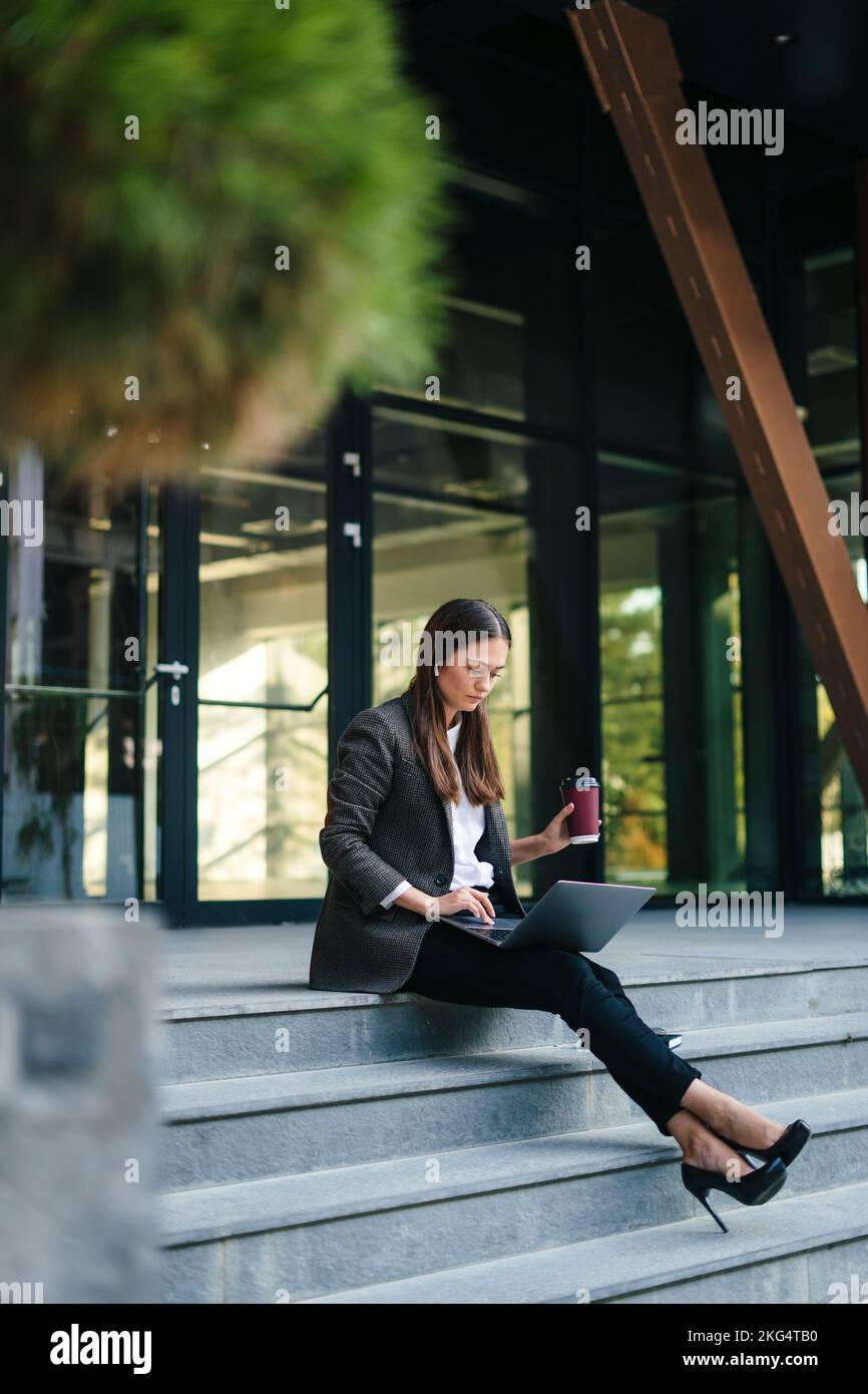 Concentrate woman specialist sitting near office building, outdoors working remote with city Wi-Fi connection. Data analysis. Business analysis. Stock Photo