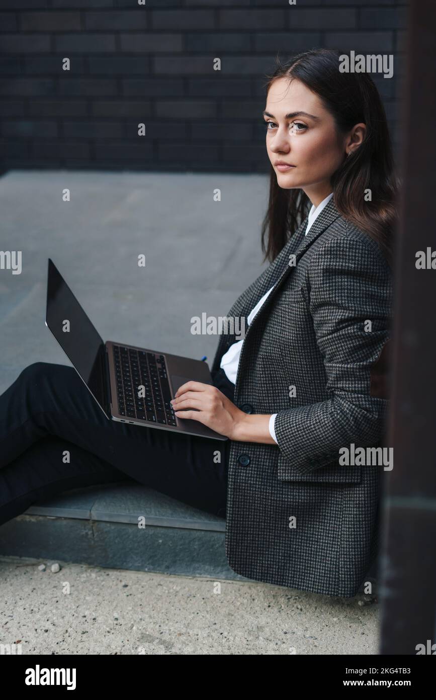 Beautiful business woman with laptop and documents, sitting on the floor leaning on the office building wall, looking away. Corporate people Stock Photo