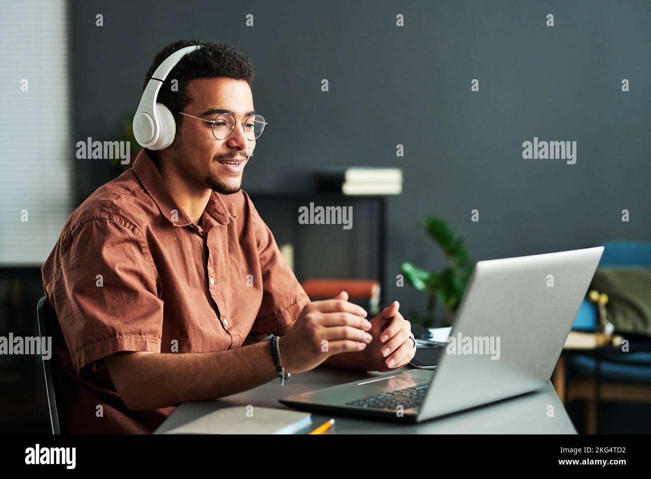 Young confident student in headphones communicating with online audience or teacher while sitting by desk in front of laptop Stock Photo