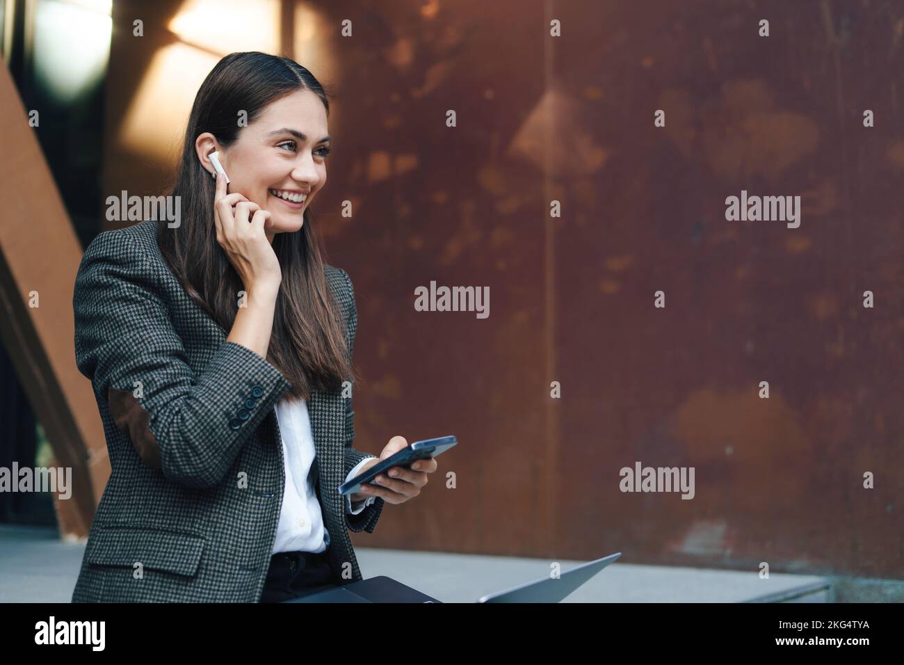 Side view of a business woman holding laptop near office outdoors, using mobile phone. Traveling, working outdoors in city, working on laptop computer Stock Photo