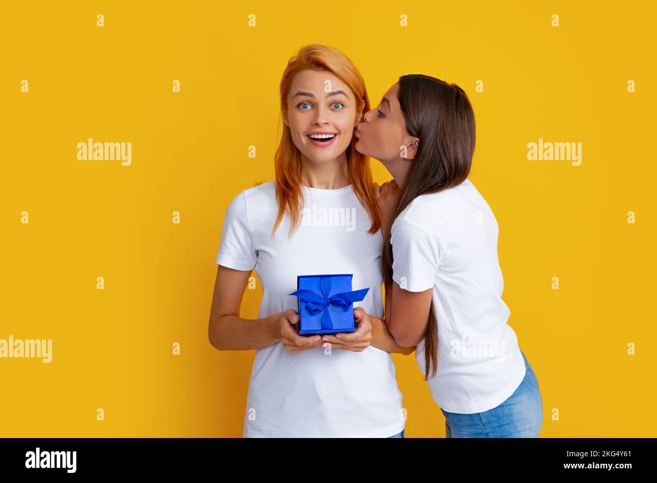 Child kissing mom. Child teen girl giving present to excited amazed mother isolated on yellow background. Holidays christmas gift. Happy mother and Stock Photo