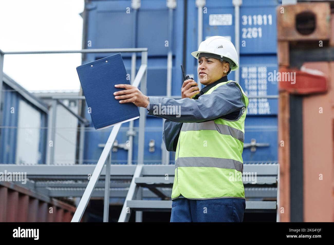 Portrait of female foreman wearing hardhat and speaking to radio while managing operations at shipping dock Stock Photo