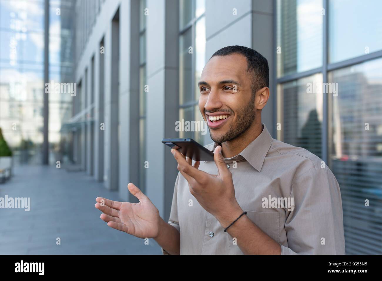 Young handsome man hispanic, african american standing outside, holding phone, talking through loudspeaker, recording on phone, smiling. Stock Photo