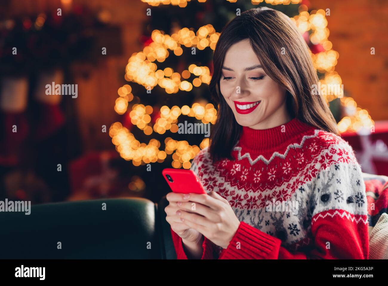 Portrait photo of young satisfied cute lady wear ugly red sweater hold her new apple iphone gift from parents chatting with friend near xmas tree Stock Photo