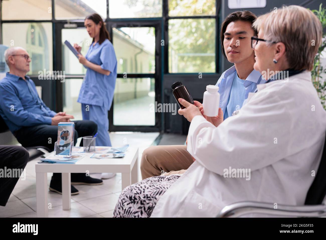 Senior medic explaining medication treatment to asian patient showing painkiller pills during checkup visit consultation in hospital room. Diverse people standing in lobby waiting to start examination Stock Photo