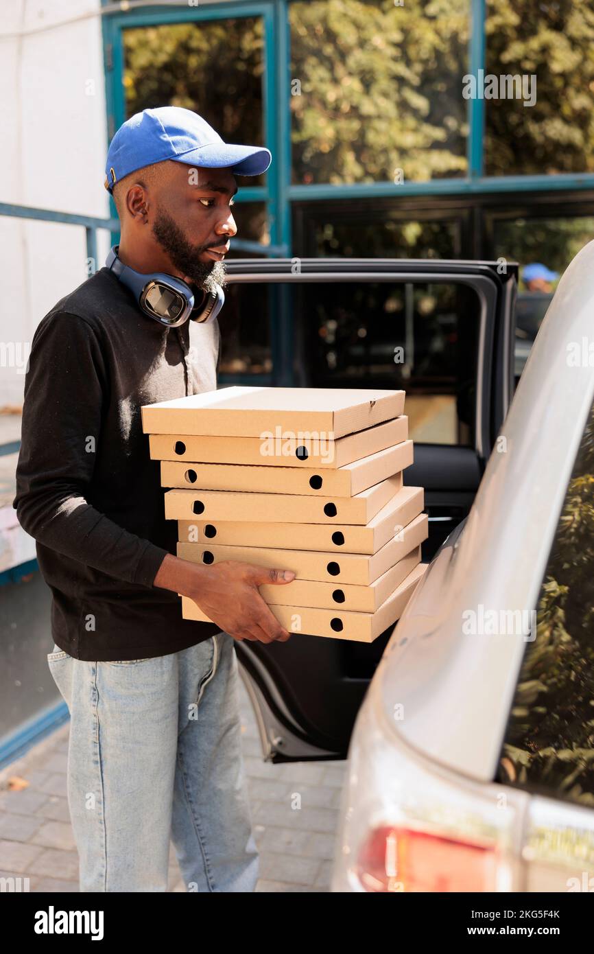 Deliveryman carrying pizza to office by car side view, african american man holding boxes pile. Lunch delivery service employee delivering fastfood, standing near company building outdoors Stock Photo