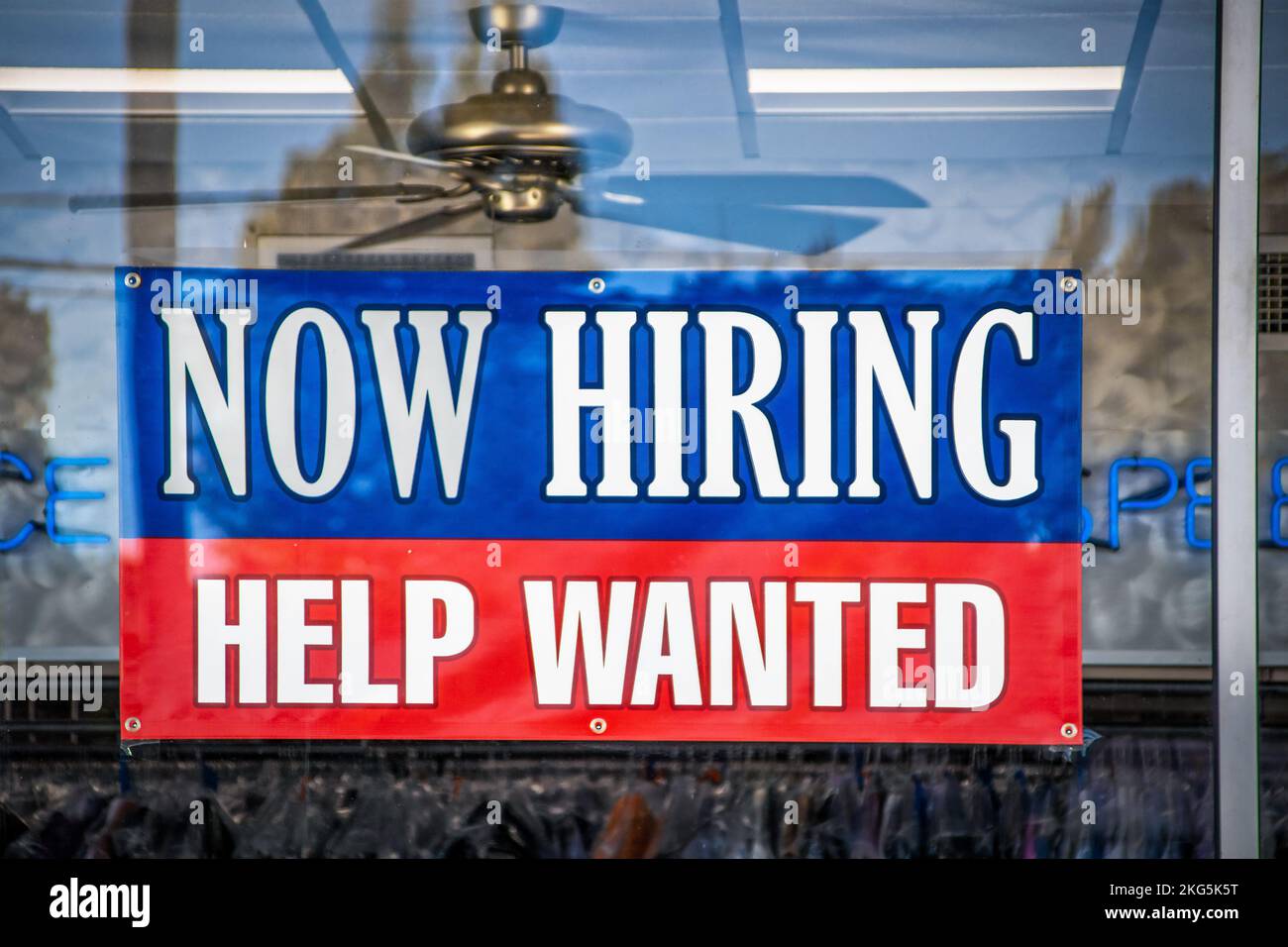 Now Hiring Help Wanted Sign in window of business with ceiling fan visible inside Stock Photo