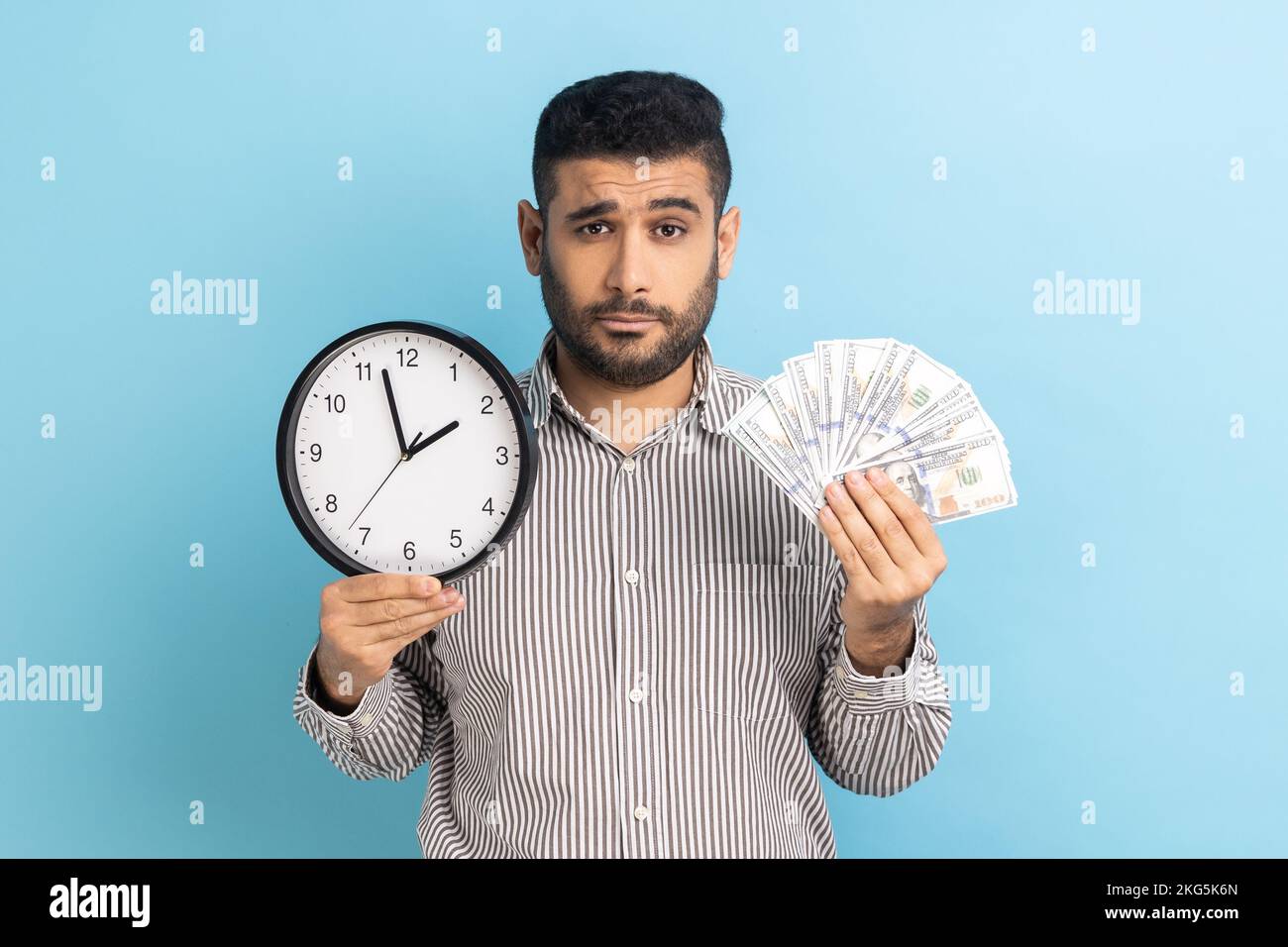 Portrait of upset sad businessman holding big fan of dollar banknotes and wall clock, time is money, looking at camera, wearing striped shirt. Indoor studio shot isolated on blue background. Stock Photo