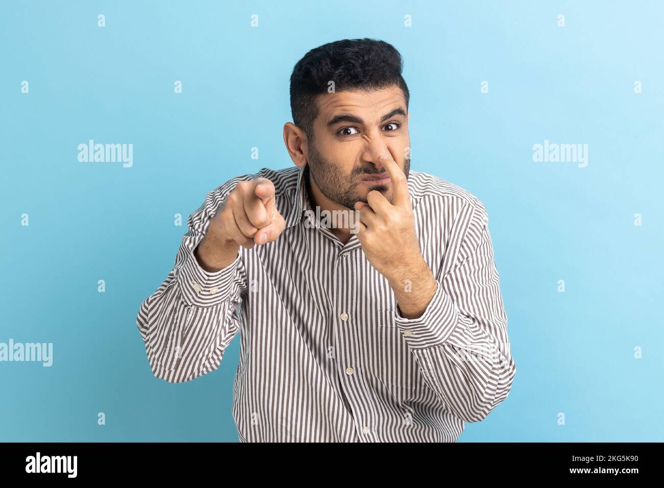 You are liar. Portrait of angry bearded man touching his nose with finger and showing lie gesture, body language, wearing striped shirt. Indoor studio shot isolated on blue background. Stock Photo