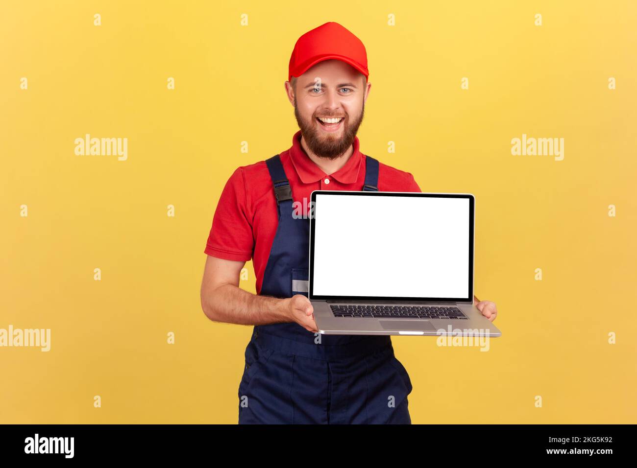 Portrait of smiling happy worker man in uniform standing and showing laptop with blank screen for advertisement, looking smiling at camera. Indoor studio shot isolated on yellow background. Stock Photo