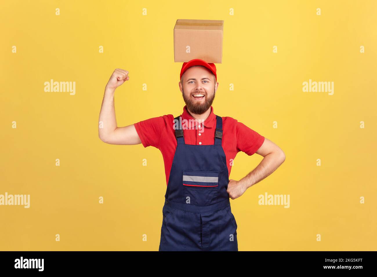 Portrait of strong courier man wearing blue uniform standing with cardboard box on his head, looking at camera, raised arm showing his power. Indoor studio shot isolated on yellow background. Stock Photo