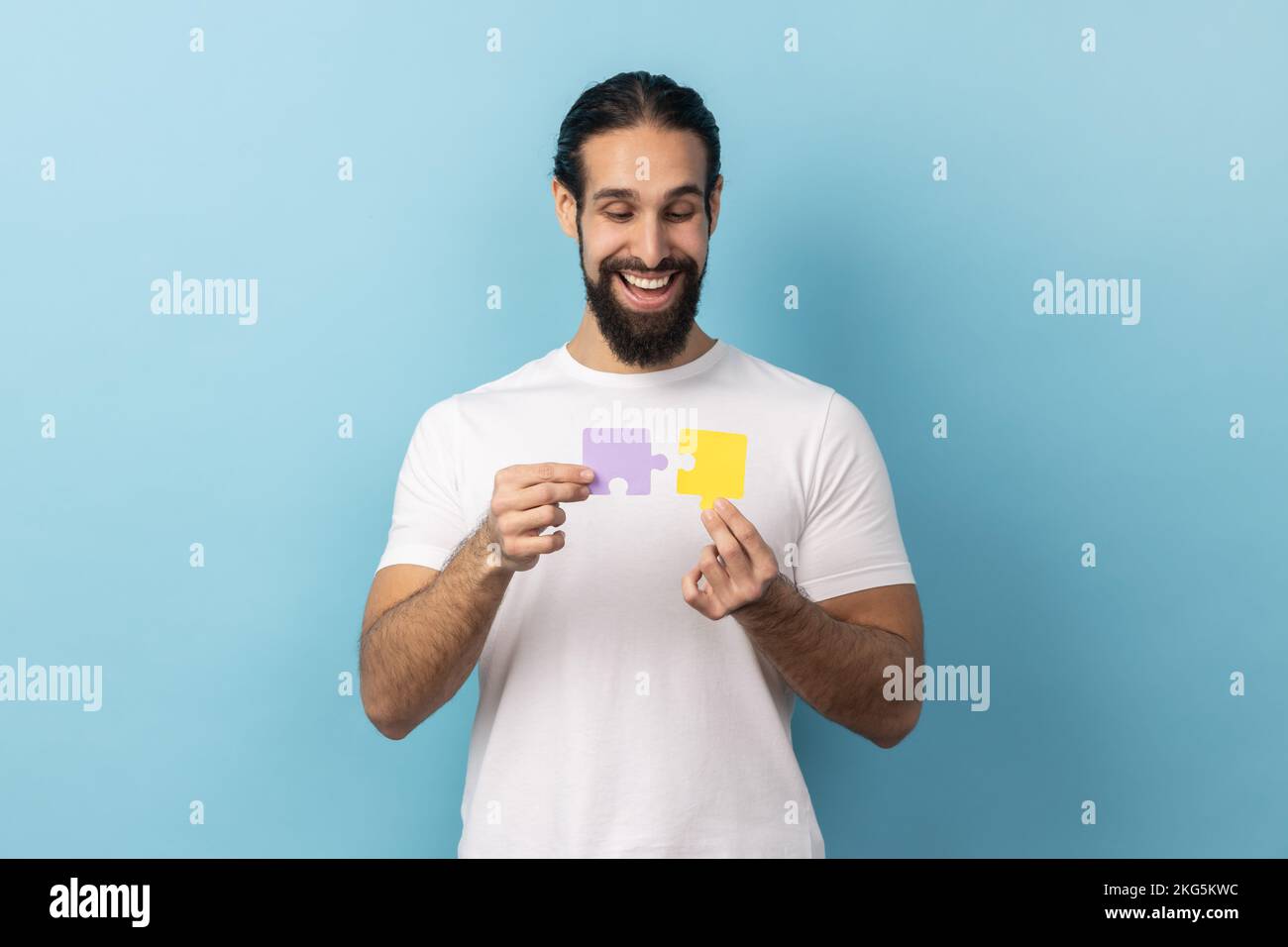 Portrait of man with beard in T-shirt holding two puzzle parts and smiling joyfully, ready to connect jigsaw pieces, symbol of union and association. Indoor studio shot isolated on blue background. Stock Photo