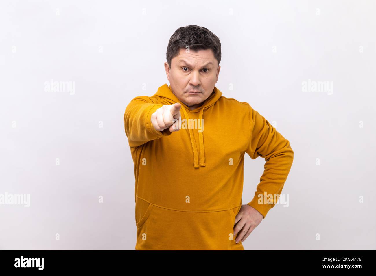 Serious middle aged man pointing finger, indicating direction to camera and looking annoyed, making choice, wearing urban style hoodie. Indoor studio shot isolated on white background. Stock Photo