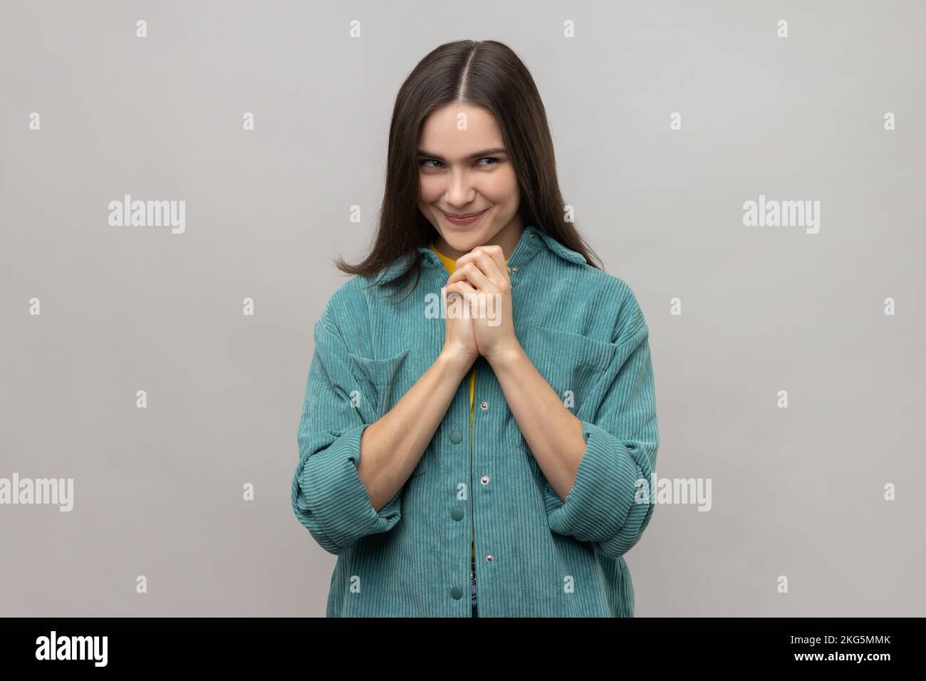 Devious cunning young woman with dark hair clasping hands and smirking mysteriously, scheming cheats, evil prank, wearing casual style jacket. Indoor studio shot isolated on gray background. Stock Photo