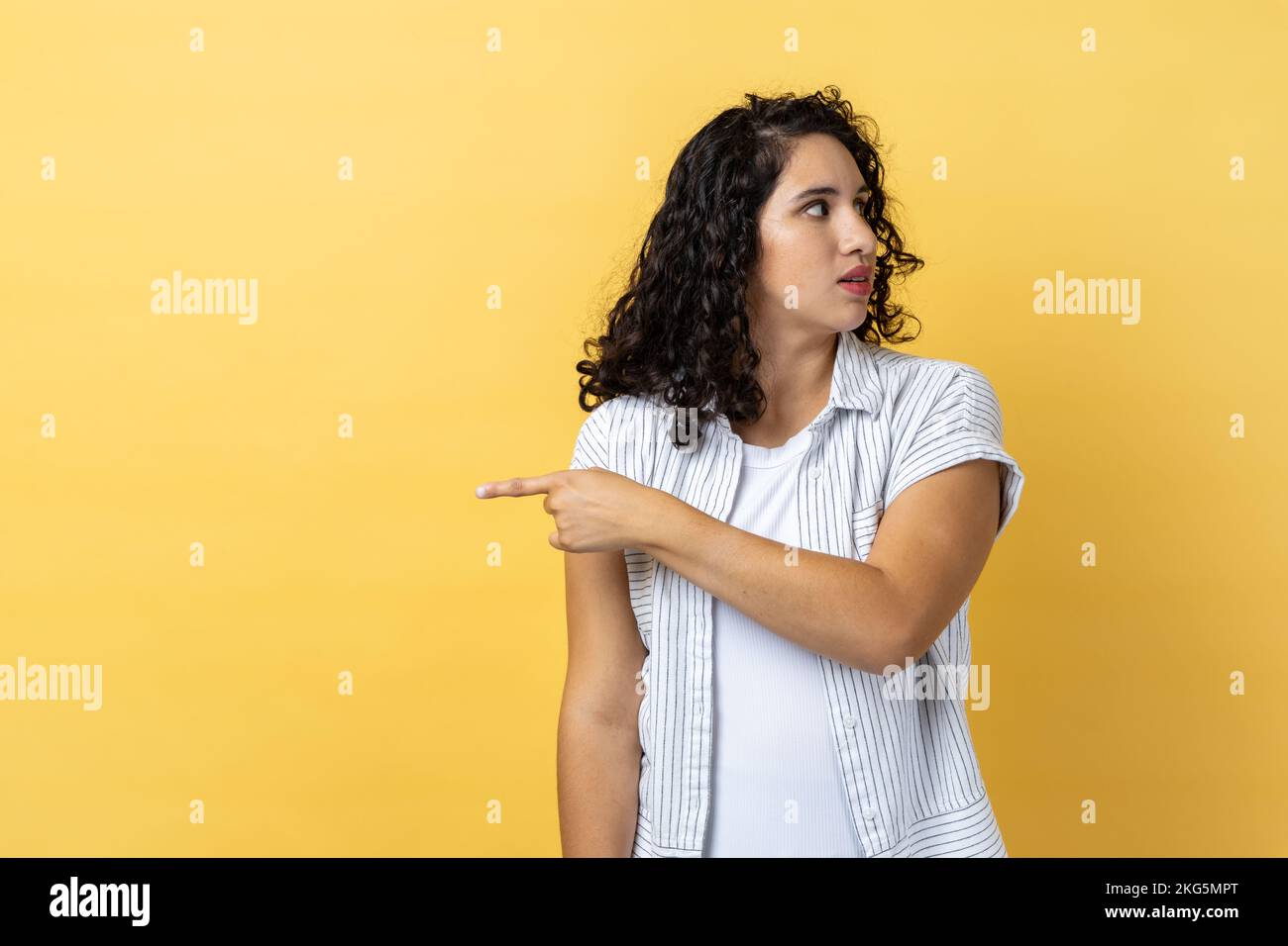 Portrait of woman with dark wavy hair pointing finger aside, ordering get out and looking resentful, boss dismissing from work, showing exit. Indoor studio shot isolated on yellow background. Stock Photo