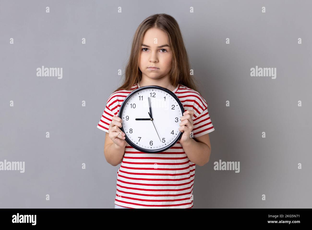 Portrait of serious little girl wearing striped T-shirt holding big wall clock, looking at camera with unpleasant emotions, time to go. Indoor studio shot isolated on gray background. Stock Photo
