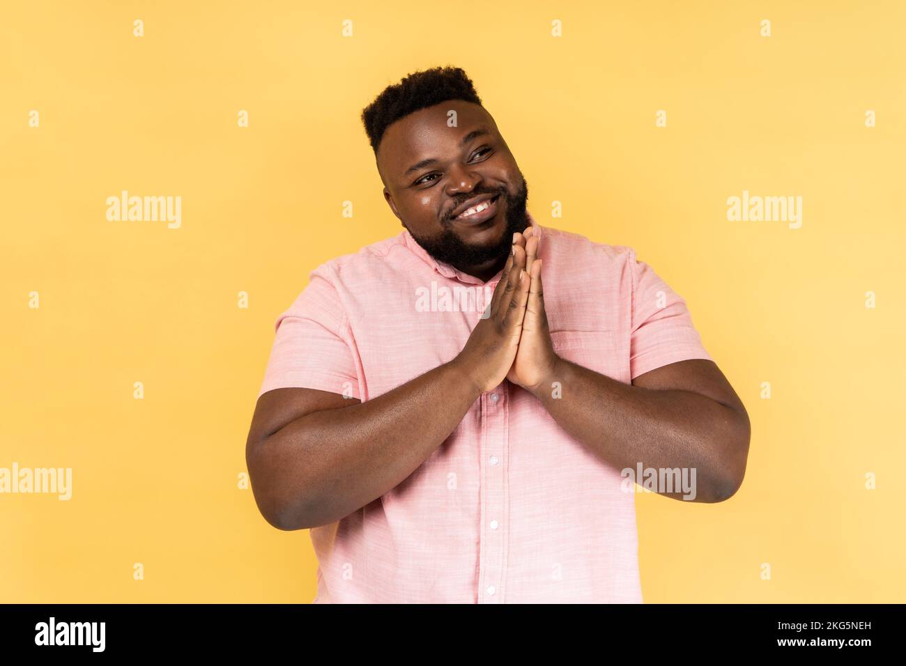 Portrait of bearded man wearing pink shirt looking away with happy expression, dreaming pleasant thoughts, keeps hands together. Indoor studio shot isolated on yellow background. Stock Photo