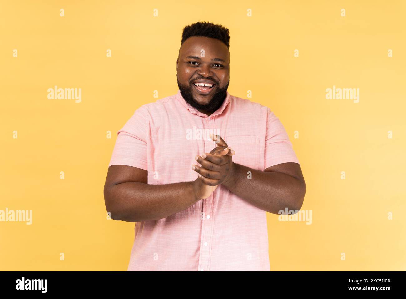 Portrait of positive friendly man wearing pink shirt standing with palms together, applauding, clapping hands congratulating you with victory. Indoor studio shot isolated on yellow background. Stock Photo