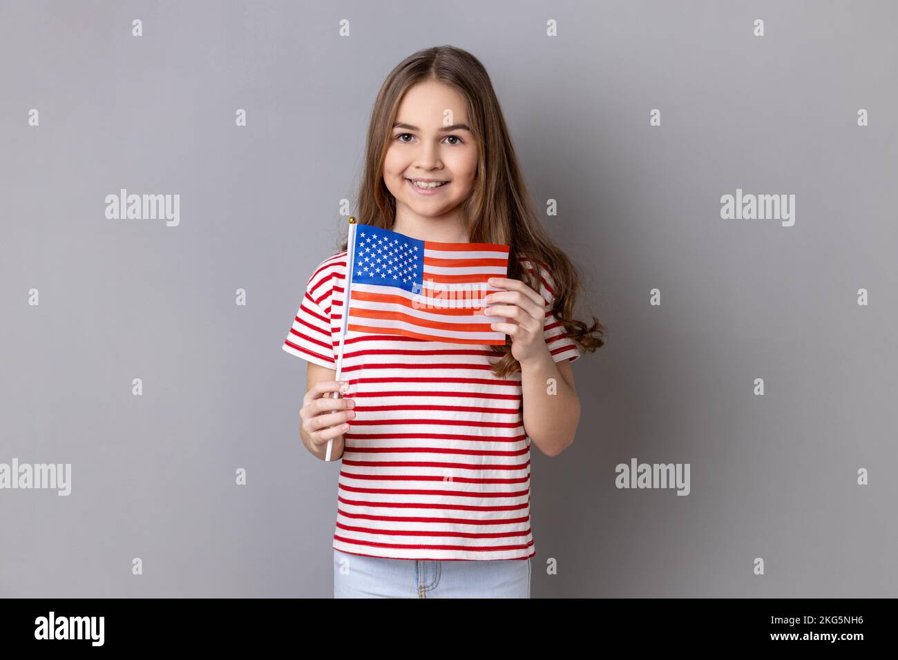 American flag. Portrait of delighted happy adorable cute little girl wearing striped T-shirt holding flag of United States of America, looking at camera. Indoor studio shot isolated on gray background Stock Photo