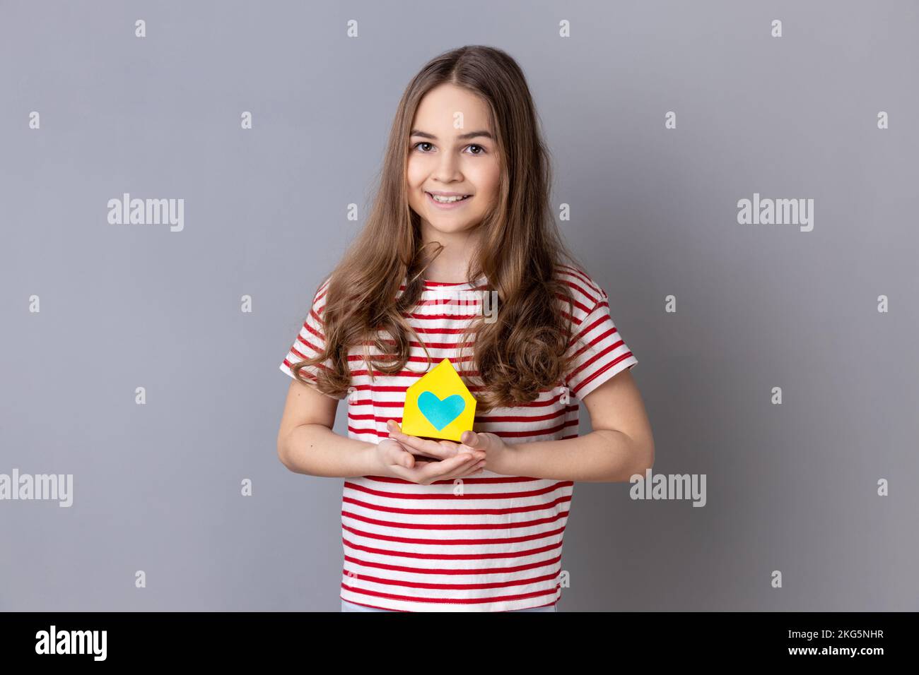 Portrait of little girl wearing striped T-shirt holding in hands little paper house looking at camera with smile, dreaming about apartment. Indoor studio shot isolated on gray background. Stock Photo