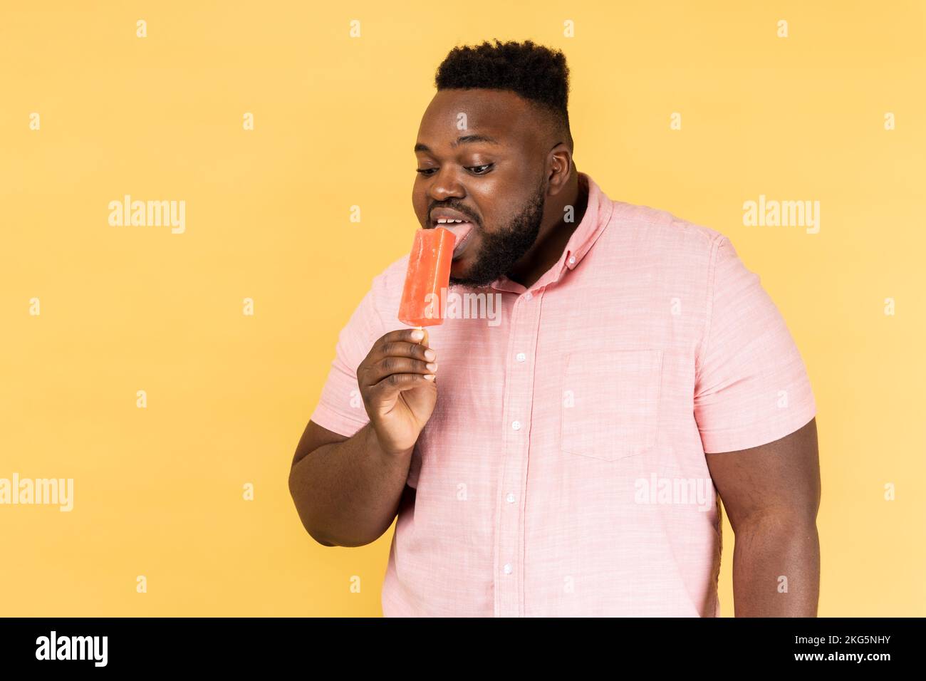 Portrait of positive optimistic man wearing pink shirt holding licking sweet ice cream, trying delicious confectionery dessert. Indoor studio shot isolated on yellow background. Stock Photo