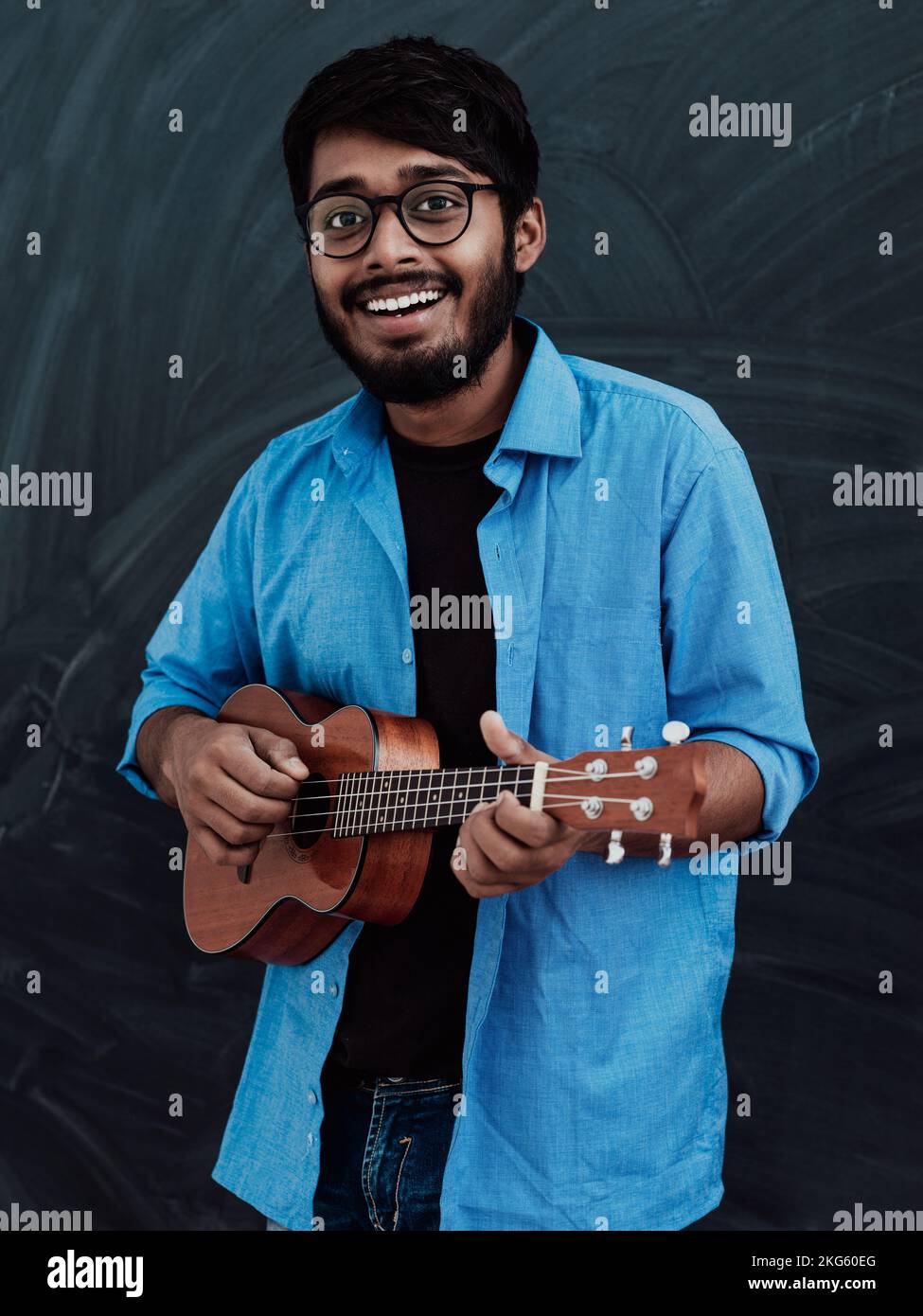 Indian young man in a blue shirt and glasses playing the guitar in front of the school blackboard Stock Photo