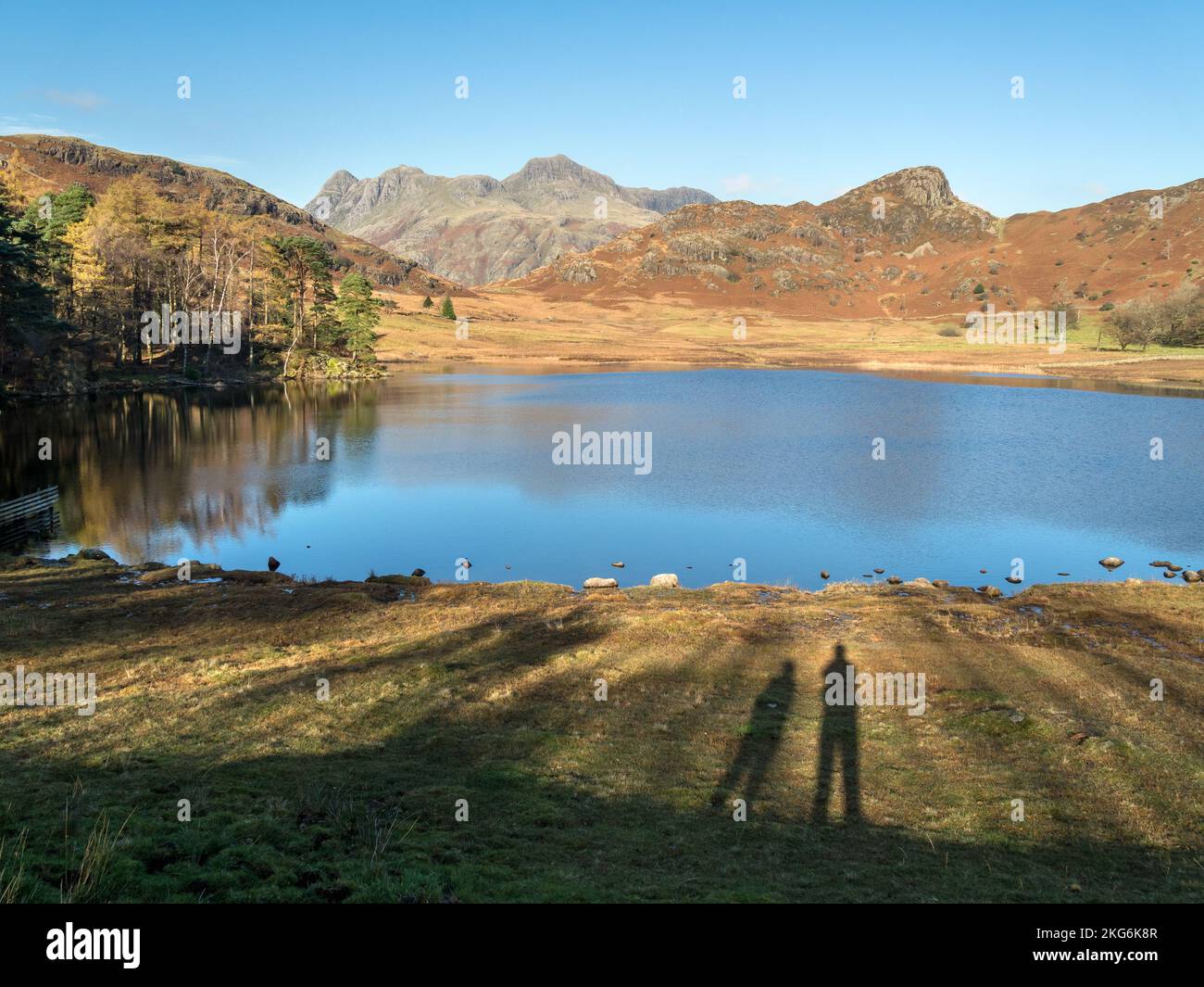 Two people casting long shadows towards Blea Tarn and the Langdale Pikes mountains in the English Lake District in Autumn, Cumbria, England, UK Stock Photo