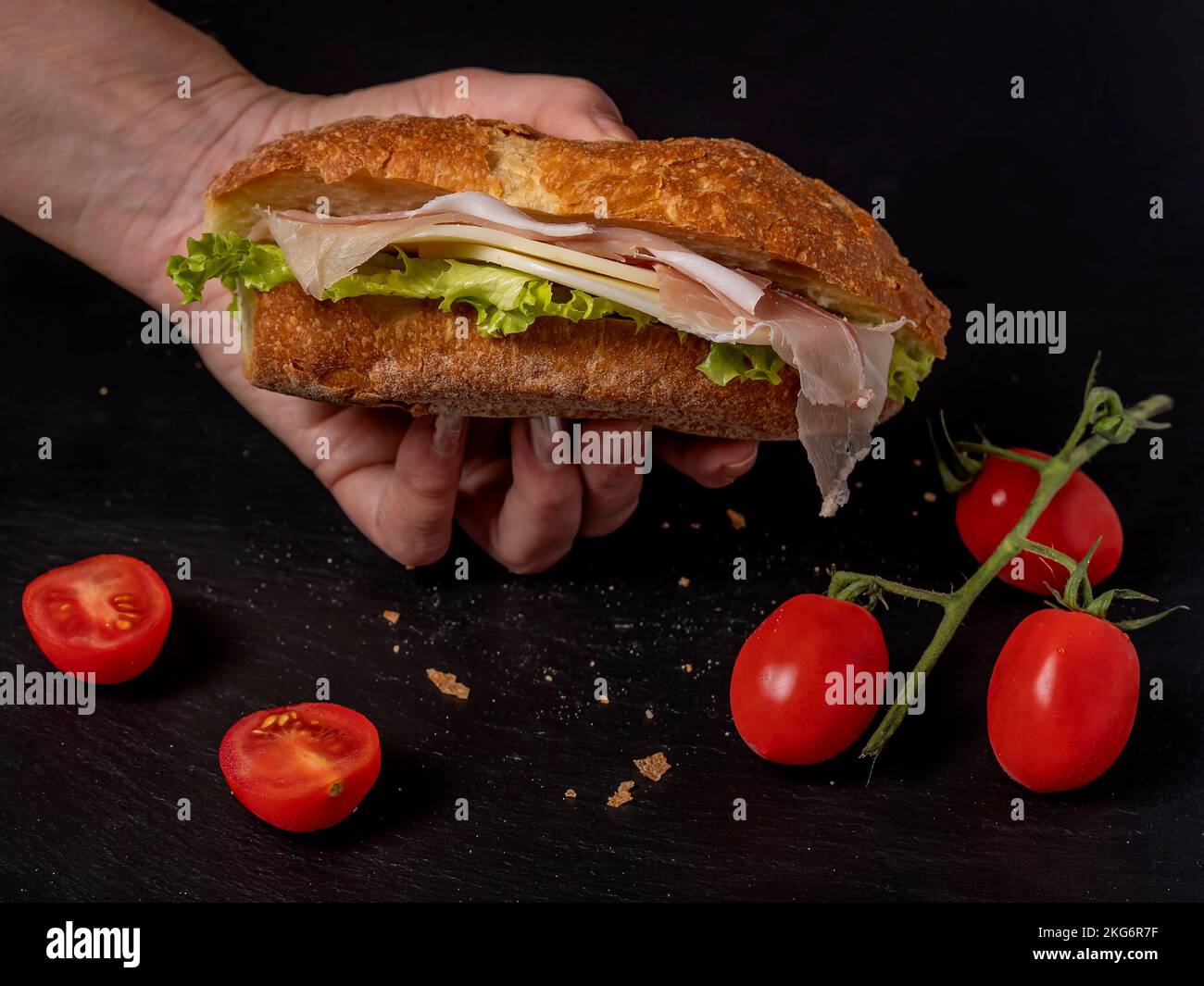 A female hand holds a sandwich filled with cheese, salad and smoked speck ham, on a black background with red cherry tomatoes Stock Photo